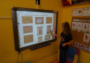 Dziewczynka stoi obok tablicy interaktywnej odszukuje z pośród różnych zegarów klepsydrę, wskazuje ją trzymanym w prawym ręku wskaźnikiem.